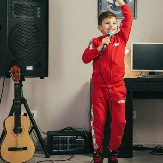 Мальчик в красном спортивном костюме поёт в комнате с музыкальной аппаратурой и гитарой