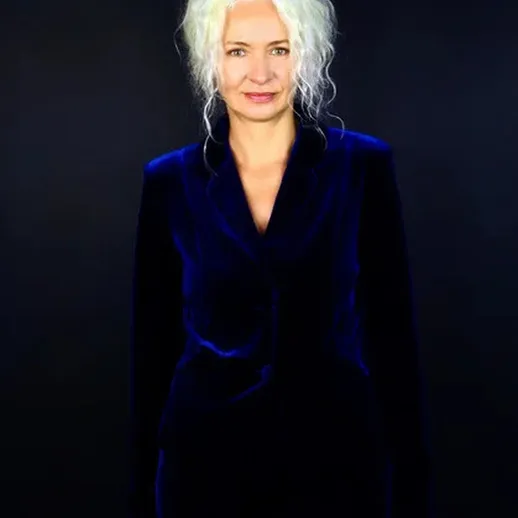 Женщина с белыми волосами в синем пиджаке на фоне темной стены в нейтральной позе