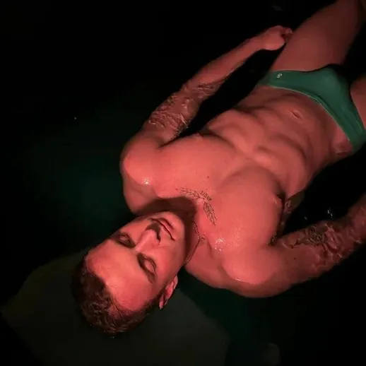 Мужчина с татуировками лежит на поверхности в темной комнате с приглушенным светом и яркими акцентами