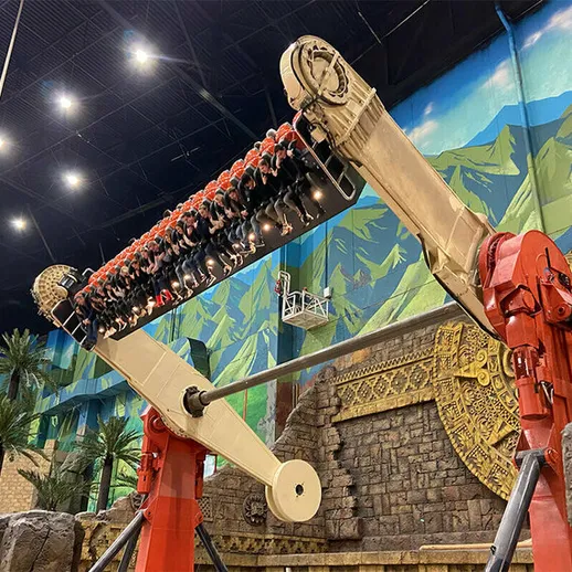 Большая аттракционная установка в развлекательном центре с ярким фоном гор и пальм, наполненная посетителями