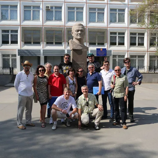 Группа людей у памятника в центре города в солнечный день, на фоне здания учебного заведения