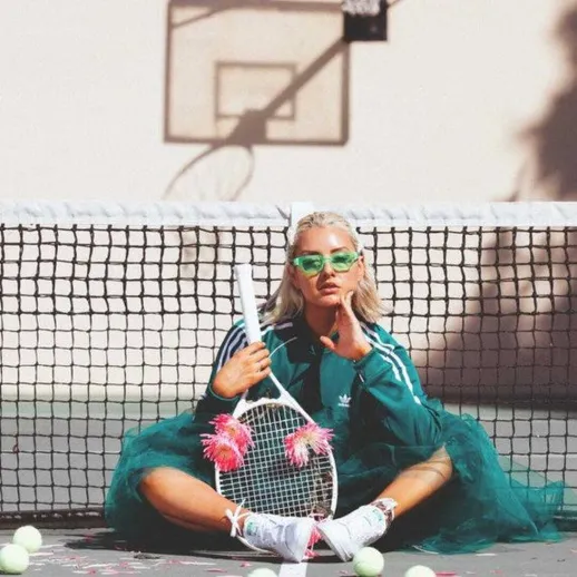 Молодая женщина в спортивной одежде и тюлевой юбке сидит на теннисной площадке с ракеткой