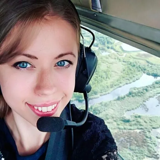 Девушка с блондинистым волосом в наушниках позирует в кабине вертолета над зелеными ландшафтами и реками