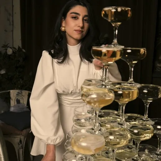 Молодая женщина в белом наряде украшает высокую пирамиду из бокалов с шампанским на праздничном мероприятии