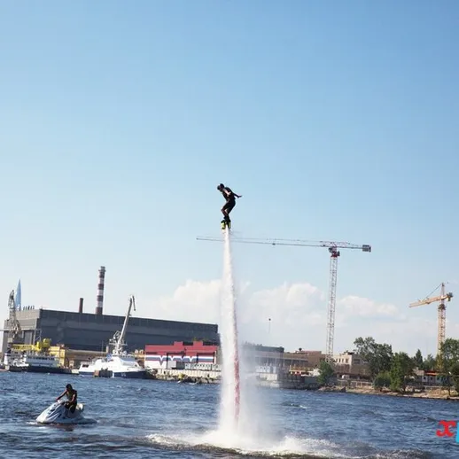 Человек на водном аппарате поднимается в воздух над рекой в солнечную погоду, на фоне городских зданий и строительных кранов