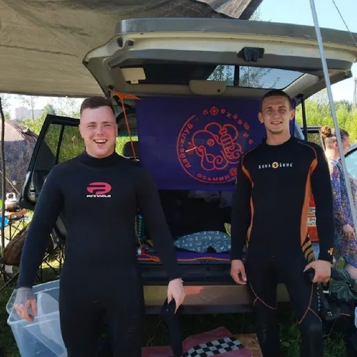 Два молодых человека в неопреновых костюмах на отдыхе у автомобиля в летний день у водоема