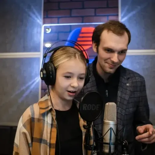 Девочка поет в студии звукозаписи, рядом с ней мужчина в пиджаке, вечернее время