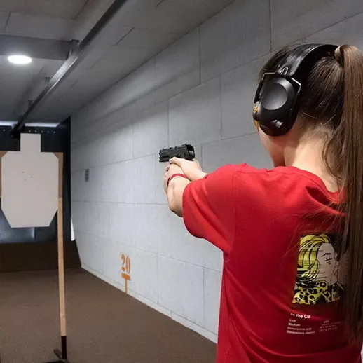 Молодая женщина тренируется в тире с пистолетом в руки, следуя указаниям инструктора и сосредоточенно целясь в мишень
