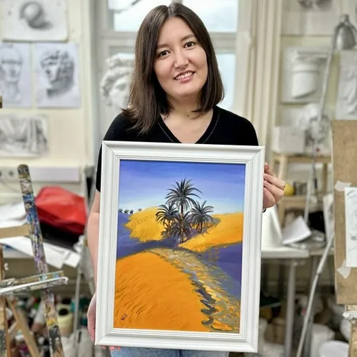 Молодая художница в ателье с картиной, изображающей пальмы на фоне пустыни