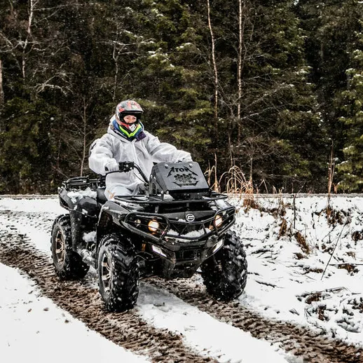 Мужчина на квадроцикле в зимнем лесу, едет по заснеженной дороге с деревьями вокруг