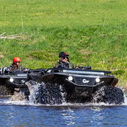 Два внедорожника перемещаются по воде в живописной местности в солнечный день с зелеными лугами на заднем плане