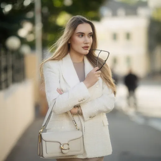 Молодая женщина в белом пиджаке прогуливается по улице с очками в руках в солнечный день
