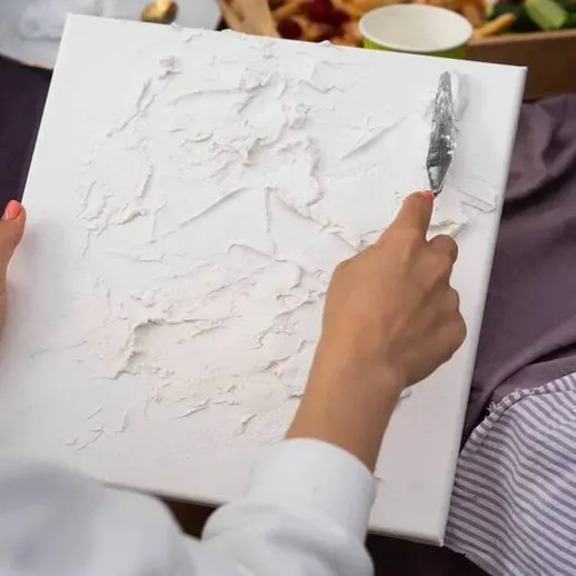 Женщина работает над художественным проектом с использованием текстурной пасты на белом холсте на фоне закусок