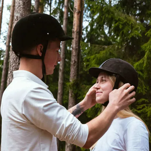 Молодой человек помогает девушке надеть шлем для верховой езды в лесу в солнечный день Молодой человек помогает девушке надеть шлем для верховой езды в лесу в солнечный день