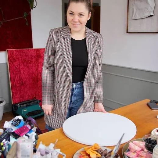 Молодая женщина в клетчатом пиджаке стоит за столом с угощениями в уютном помещении творческой студии