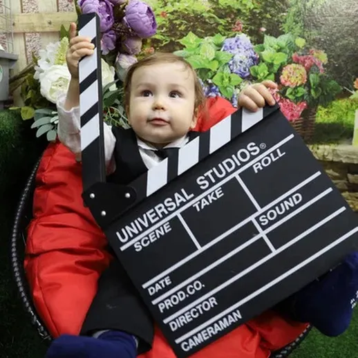 Младенец в костюме держит киносценарный клипс в декорациях на фоне цветов