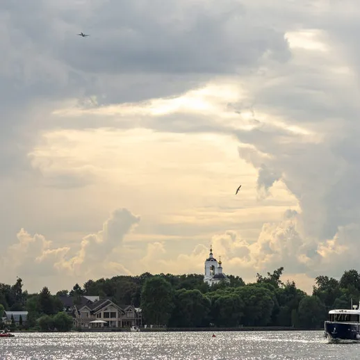 На горизонте виден маяк и выходящие на берег облака в лицо заката на спокойной воде реки