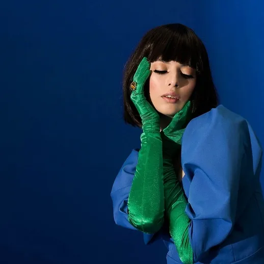 Модель в синем платье и зелёных перчатках позирует на фоне яркой стены