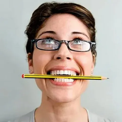 Женщина улыбается с карандашом в зубах на сером фоне, демонстрируя креативный подход к обучению и работе