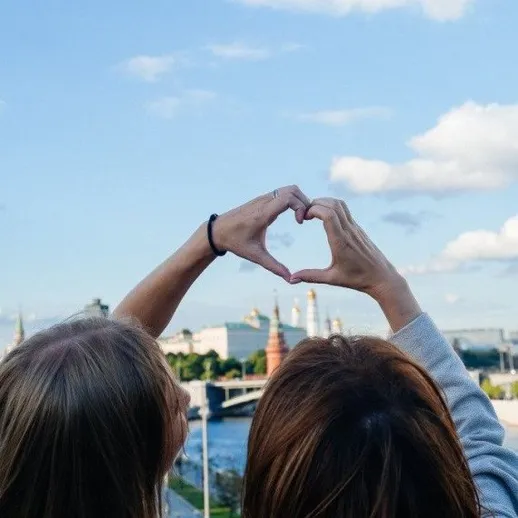 Две женщины показывают сердечко на фоне города и реки в солнечный день
