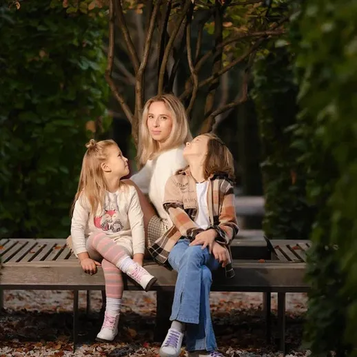 Мать с двумя дочерьми на скамейке в парке осенью, окружённые зеленью и цветами, в спокойной обстановке