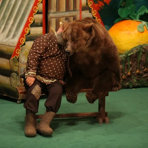 Человек и медведь сидят на сцене в театре, окруженные декорациями на фоне ярких растений и цветов