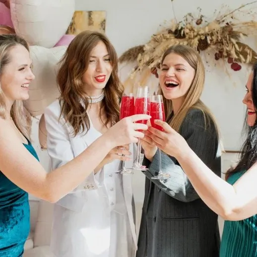 Четыре женщины радуются на вечеринке, поднимая бокалы с напитками в ярком интерьере