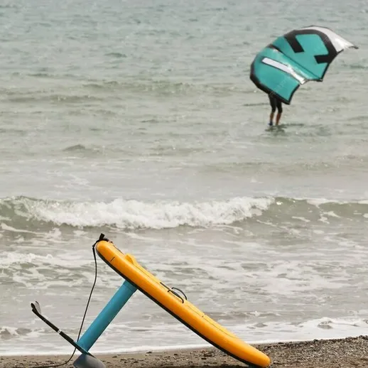 На пляже в летний день лежит кайт и доска для кайтсерфинга рядом с катающимся серфером на волнах