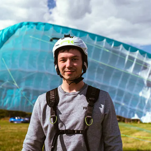 Парапланерист в шлеме улыбается на лужайке с парапланом под ярким голубым небом