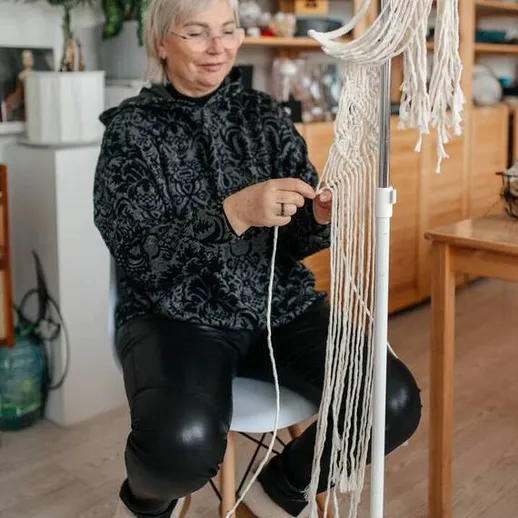 Женщина работает над макраме в уютной мастерской, окруженной растениями и рукодельными изделиями