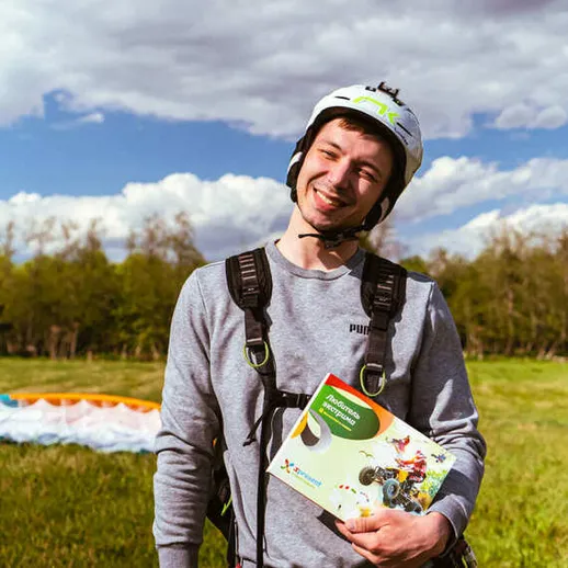 Парапланерист в спортивной экипировке на фоне зеленого поля и облачного неба