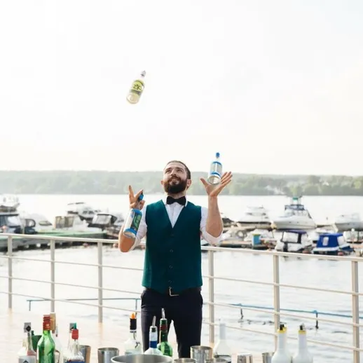 Бармен выполняет жонглирование бутылками на открытой террасе у реки во время солнечного дня