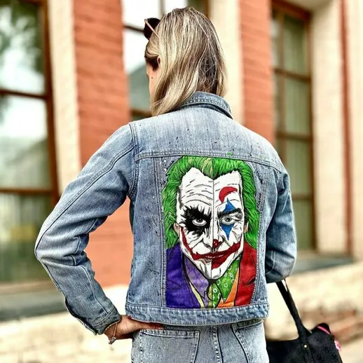 Молодая женщина в джинсовой куртке с изображением Джокера на спине прогуливается по улице в ясный день