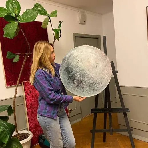 Молодая женщина держит декоративное изображение луны в уютном интерьере мастерской на фоне растений и художественных элементов