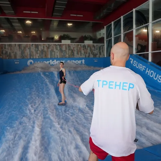 Молодая девушка обучается серфингу на искусственной волне в спортзале, наблюдающий инструктор помогает ей с балансом