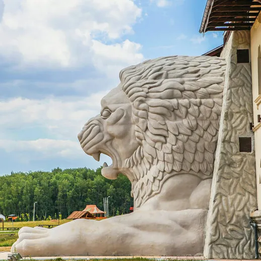 Огромный каменный лев у здания, расположенного на фоне зелёных лесов в ясный день