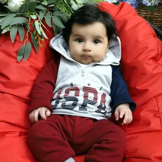 Младенец в ярком спортивном костюме сидит на уютном кресле во дворе среди зелени в солнечный день