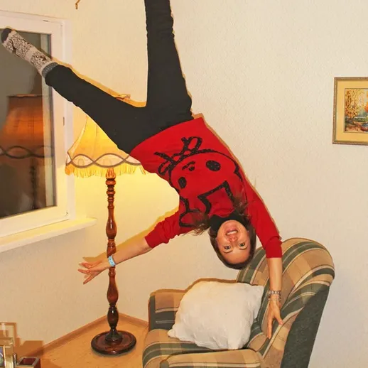 Молодая женщина выполняет акробатический трюк, висит вниз головой в уютной комнате с диваном и лампой