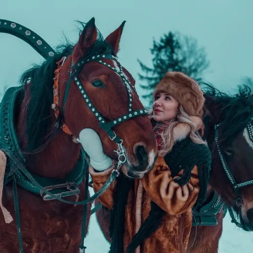 Женщина ласкает лошадей на зимней ферме среди снежного пейзажа в облачном свете