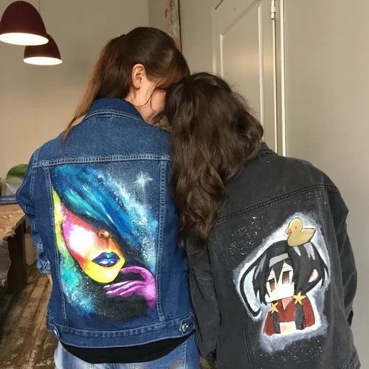 Девушки в джинсовых куртках с яркими рисунками стоят вдвоем в интерьере, украшенном стильными светильниками