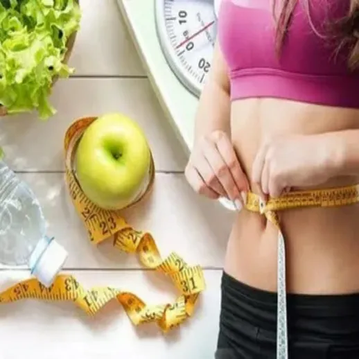 Женщина измеряет свою талию на кухне, рядом с фруктами и бутылкой воды