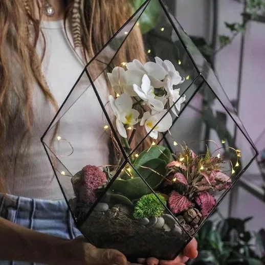 Женщина с террариумом в руках, украшенным орхидеями, в интерьере с зелеными растениями и огоньками