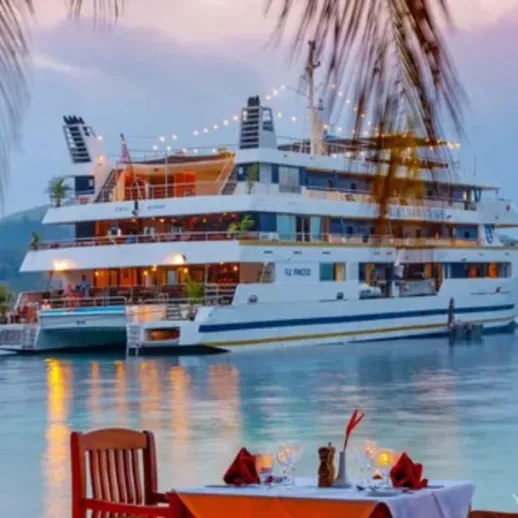 Круизный корабль у берега на закате, пальмы и столики для ужина на берегу