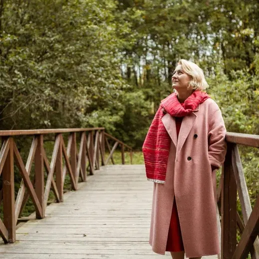 Женщина в розовом пальто стоит на деревянном мостике среди зелёных деревьев в солнечный день