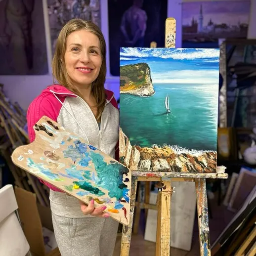 Женщина художник с картиной морского пейзажа и палитрой в мастерской, светлый интерьер, вечернее время