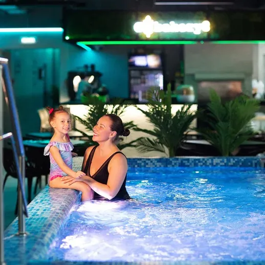 Женщина сидит на краю бассейна и играет с девочкой в помещении с освещением