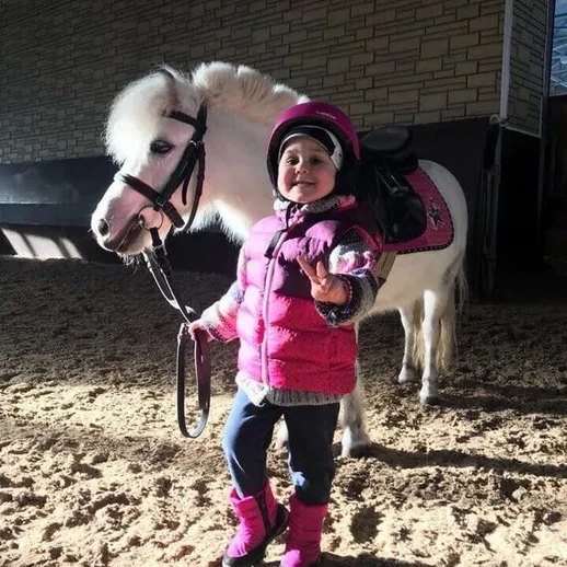 Девочка на пони в зимней одежде в конюшне, счастливо позирующая с улыбкой