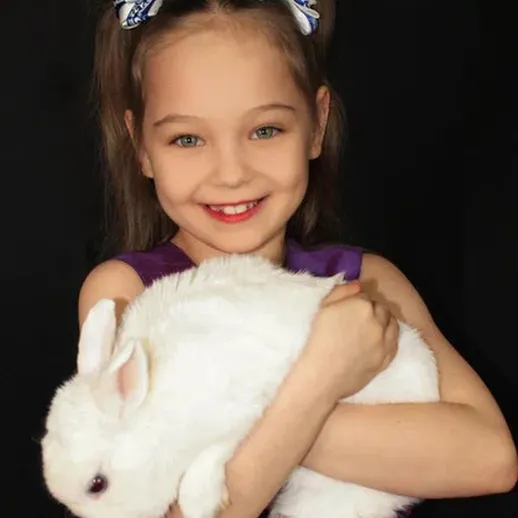 Девочка с белым кроликом на черном фоне, улыбается и обнимает его в ярком платье