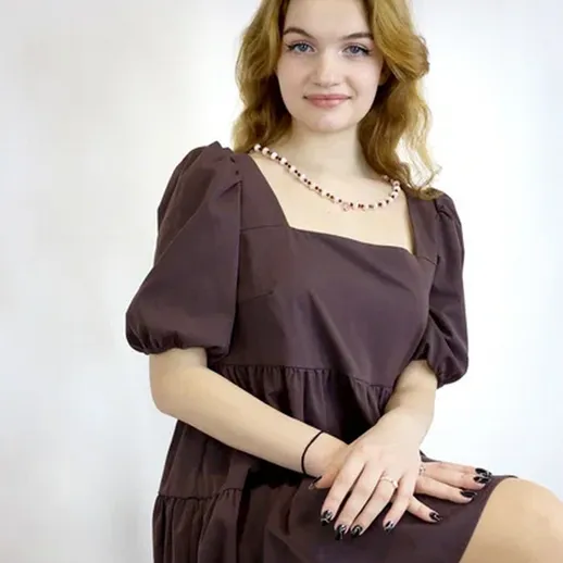 Молодая женщина в темном платье позирует на нейтральном фоне с легкой улыбкой и украшением на шее