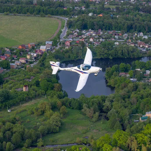 Летящий легкий самолет над зелеными полями и рекой в тихом пригороде, указывая на ясное небо в дневное время